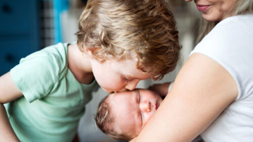 Male toddler kissing infant sibling being held in mother's arms