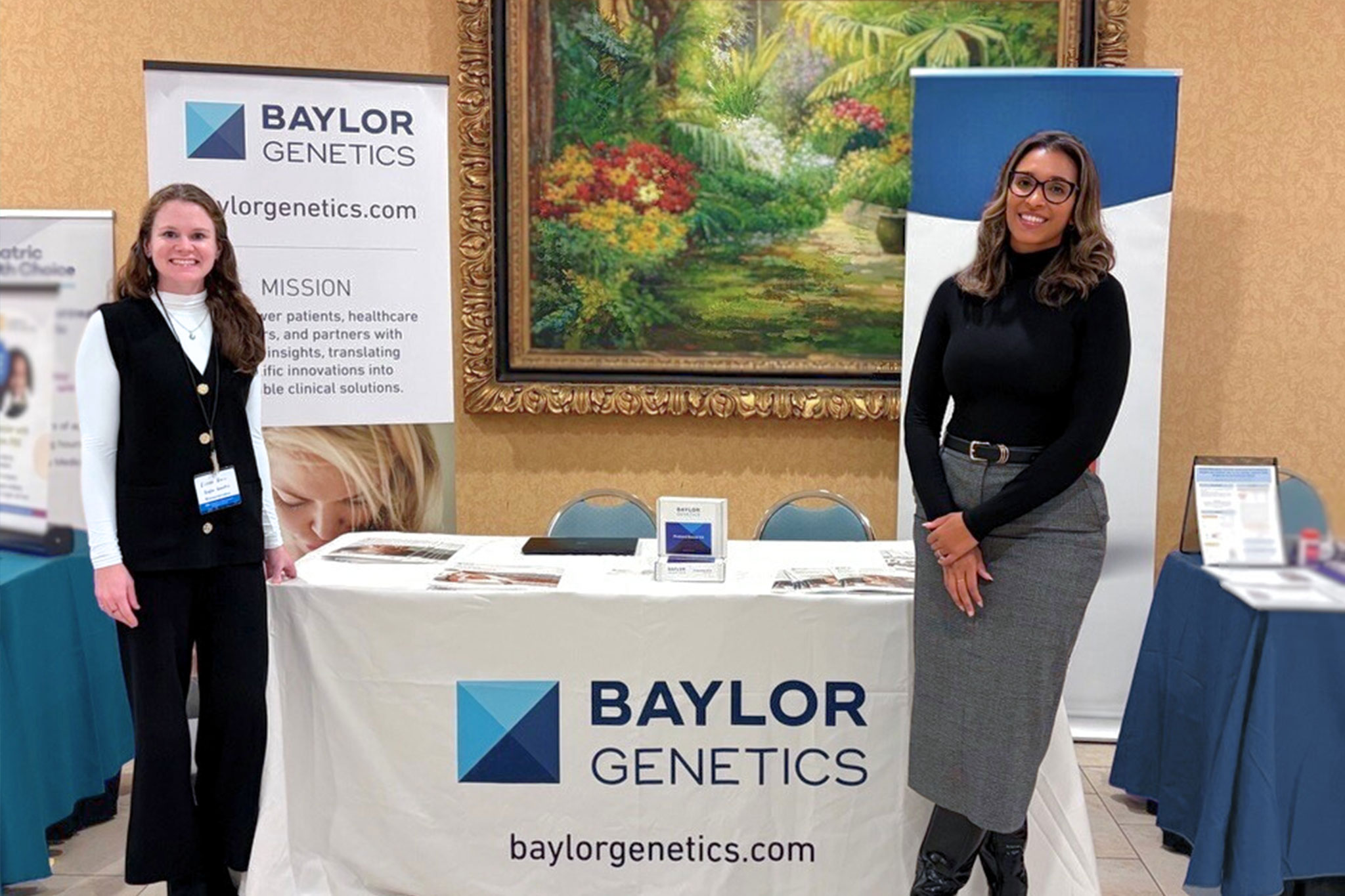 Two female Baylor Genetics employees standing in front of Baylor Genetics table and marketing material